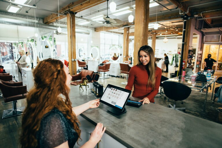 Dans un salon de coiffure, une cliente et une vendeuse discutent à la caisse dans une bonne ambiance.